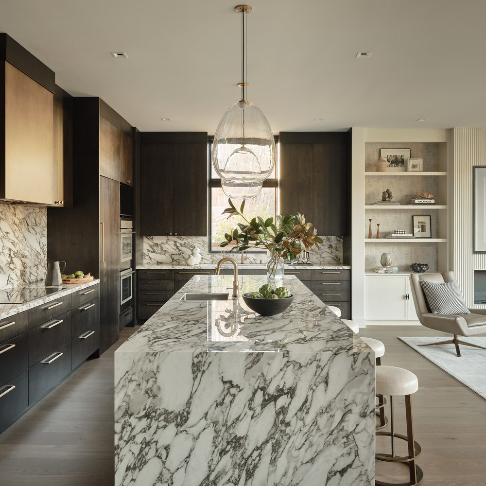 Bel Aire kitchen featuring dark stained oak cabinetry, a veined marble waterfall island and full-height backsplash, antique brass fixtures, and built-in shelving beside a light-filled living area