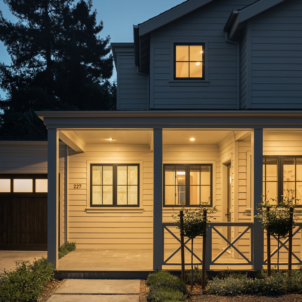 Sycamore Park custom modern farmhouse in Mill Valley featuring white horizontal siding, black-framed windows, covered front porch with X-pattern railings, open-concept interior with light oak floors, white shaker kitchen with waterfall island, and indoor-outdoor living spaces with folding glass doors
